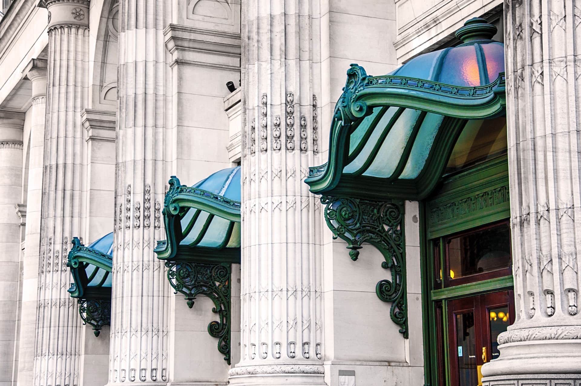 Tres marquesinas de bronce con pátina verde y cúpulas de vidrio azul, entre columnas acanaladas de piedra gris de una fachada clásica.