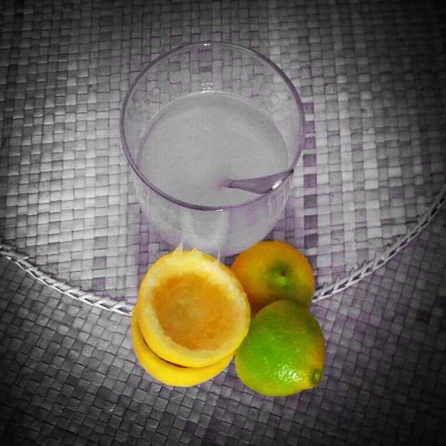 Vaso de agua con una cuchara dentro, junto a medio limón, un limón entero y una lima, sobre un mantel de rejilla gris.