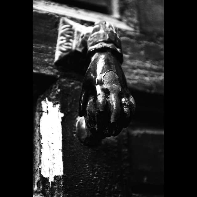 Aldaba de hierro con forma de mano cerrada sobre una puerta oscura, en blanco y negro y con fuerte contraste.