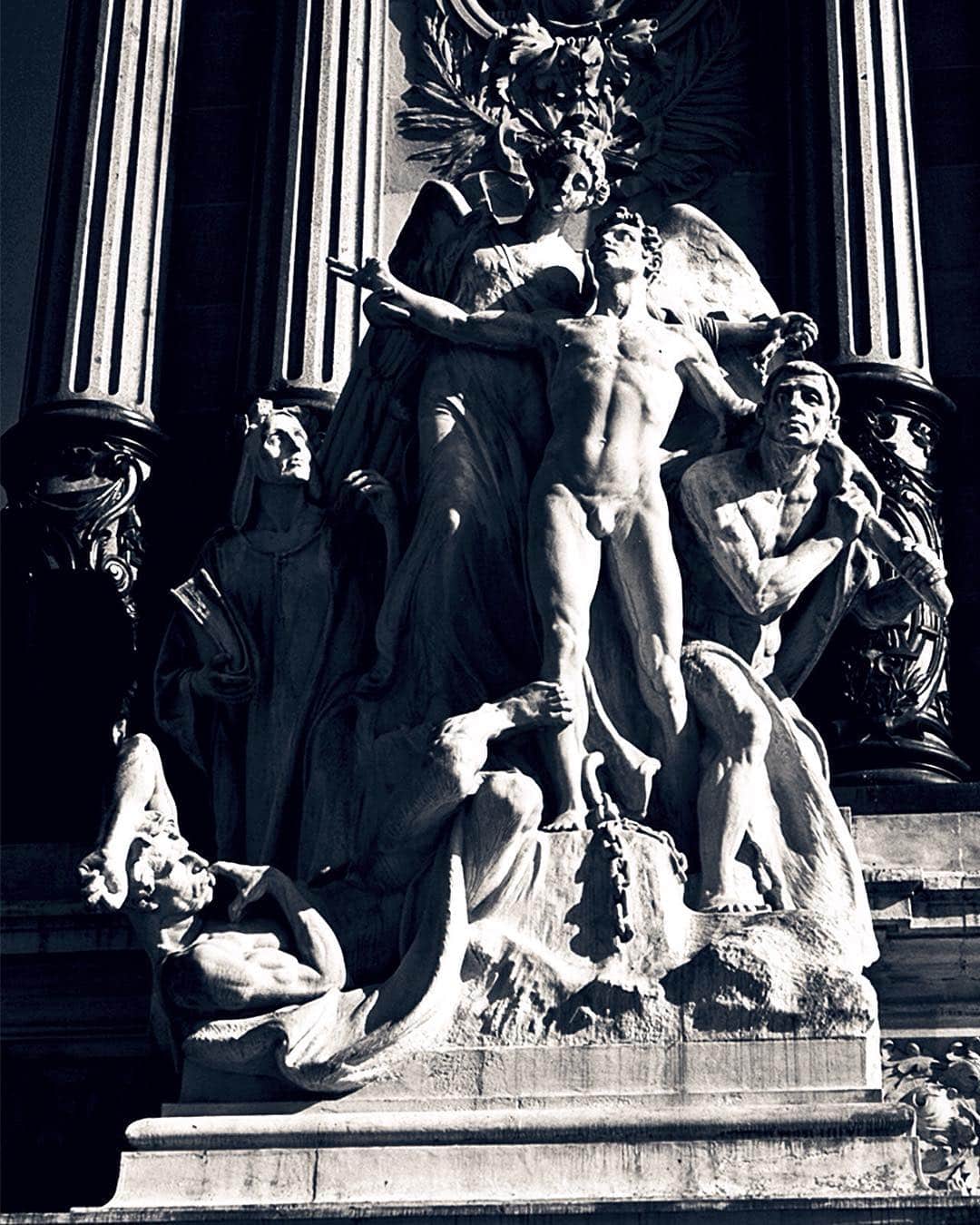 Grupo escultórico de mármol con figuras desnudas y un ángel, al pie de unas columnas, en blanco y negro.