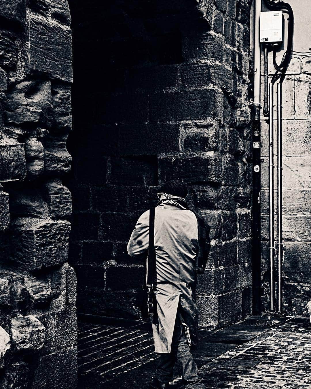 Hombre de espalda subiendo unas escaleras estrechas entre muros de piedra, en blanco y negro.
