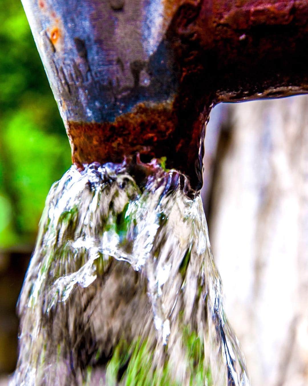 Chorro de agua saliendo a presión de un caño de hierro oxidado, con el fondo verde desenfocado.