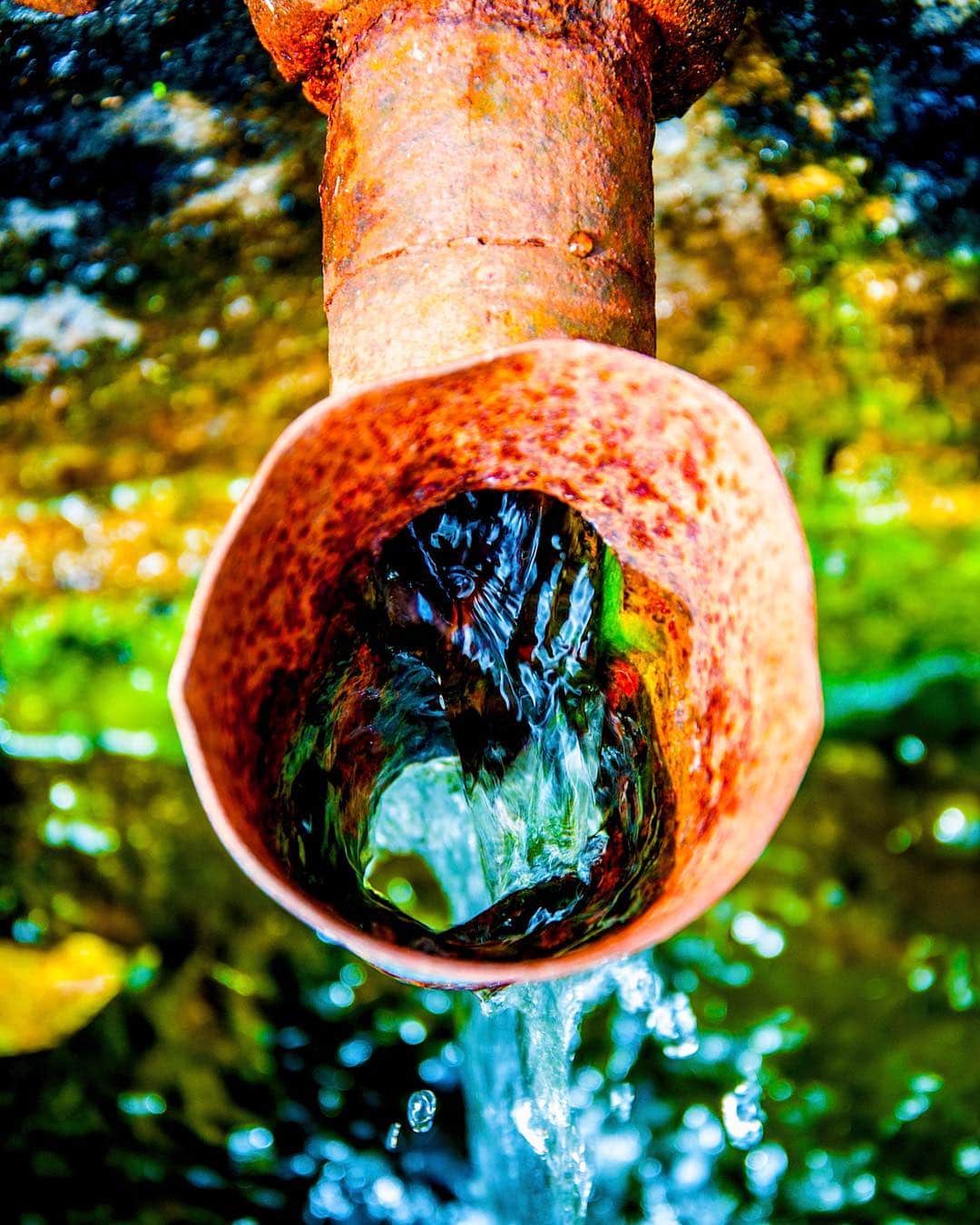 Boca de un caño oxidado en primer plano, con el agua saliendo y el follaje otoñal desenfocado detrás.