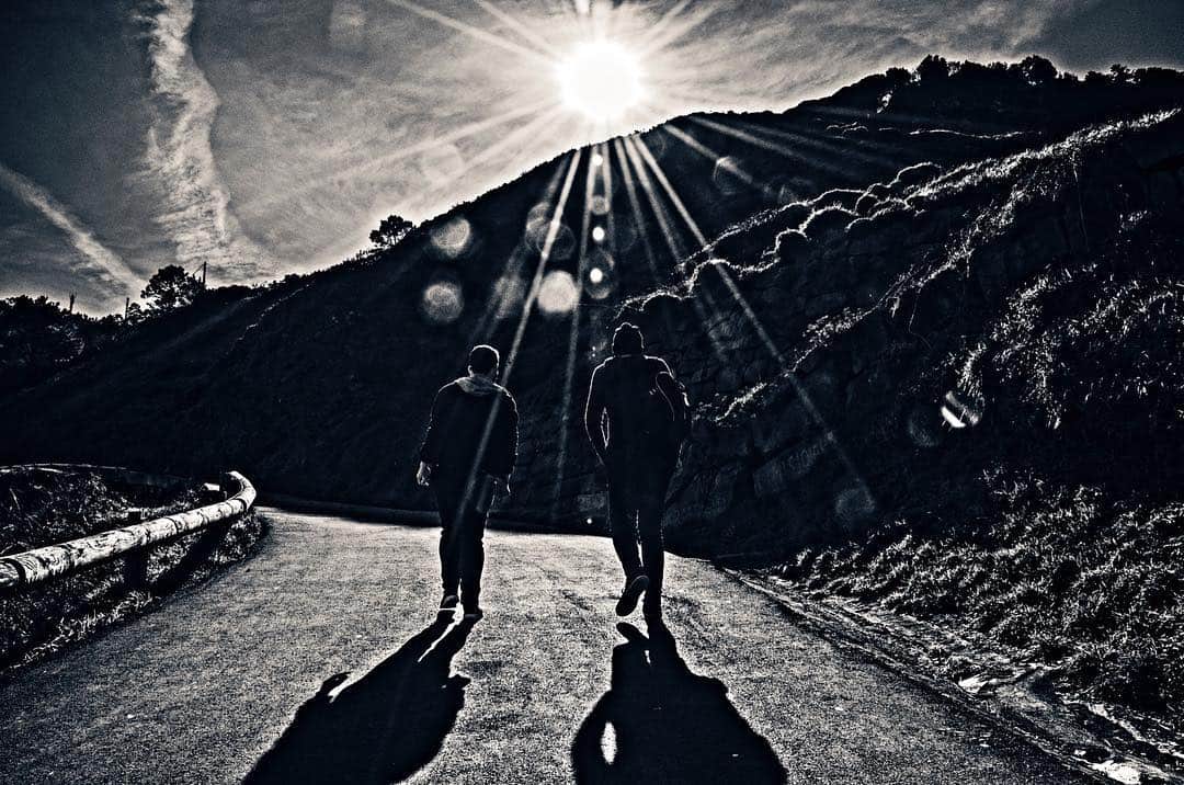 Dos siluetas caminando cuesta arriba por una carretera a contraluz, con el sol en estrella, en blanco y negro.