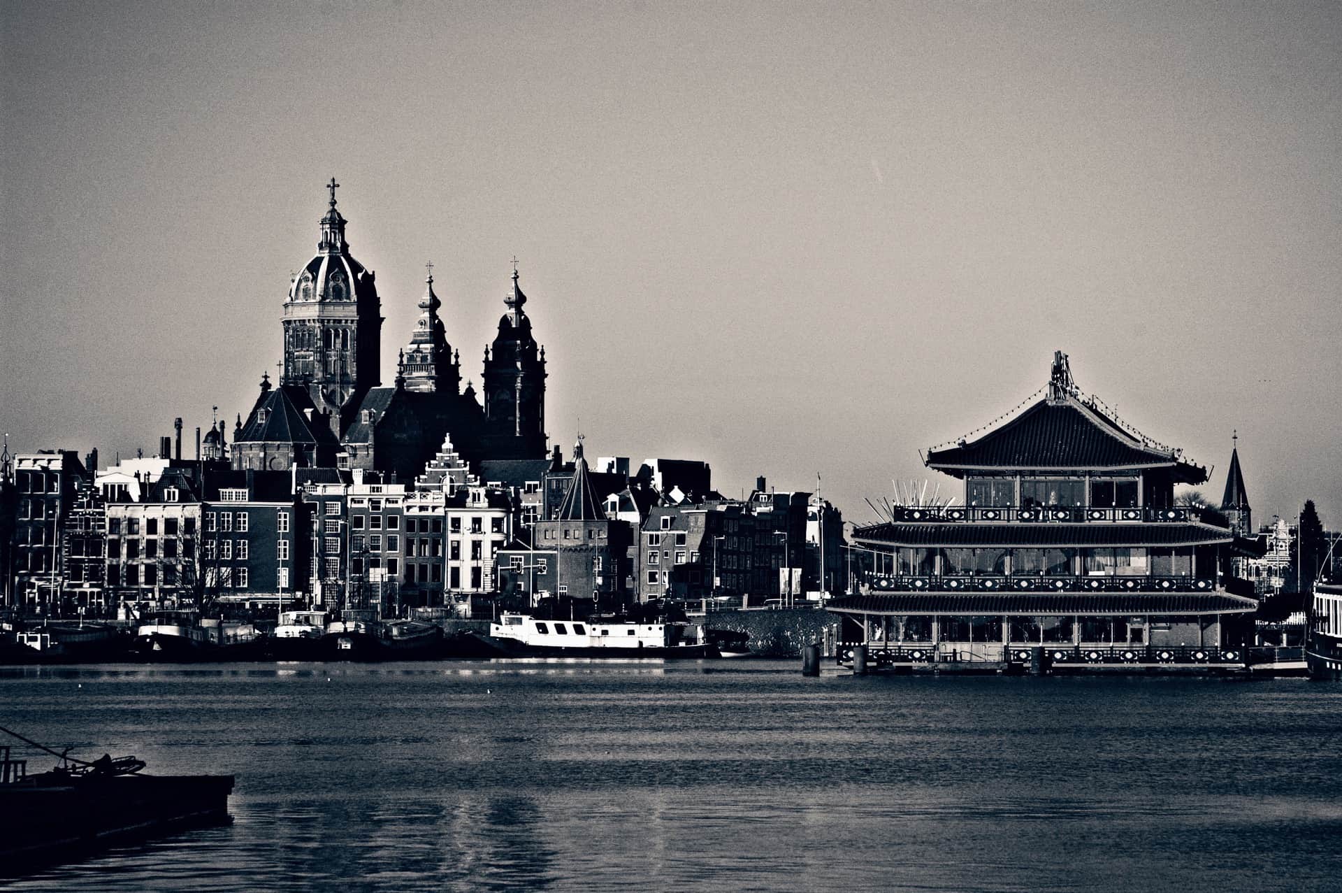 Perfil urbano al otro lado del agua, en blanco y negro: casas con hastiales, iglesia de cúpula y un edificio flotante con tejados chinos.