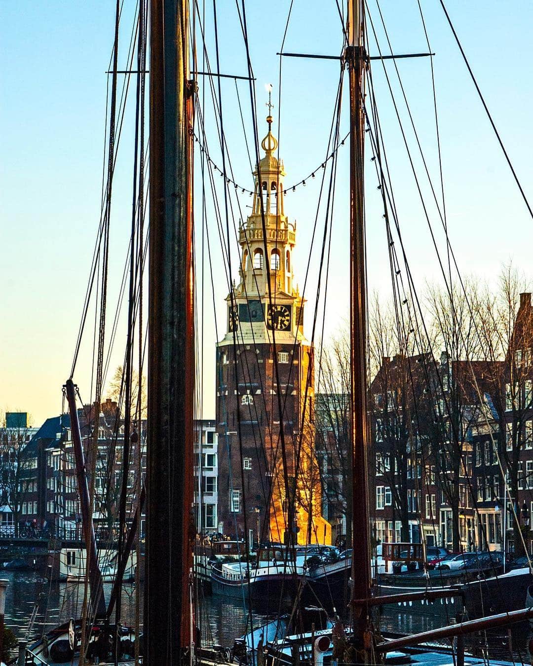 Mástiles de barcos amarrados en un canal de Ámsterdam, con una torre dorada al fondo al atardecer.