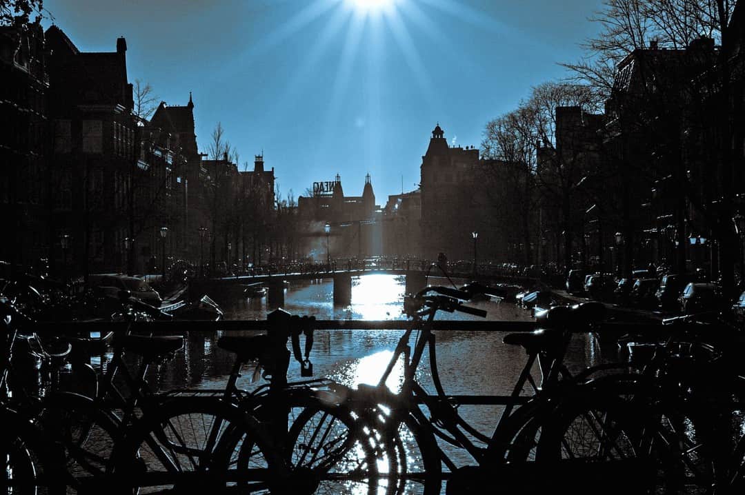 Canal de Ámsterdam a contraluz con el sol en estrella, bicicletas apoyadas en el pretil y el agua brillando.
