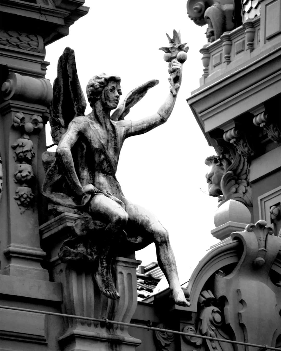 Escultura de un ángel sentado sobre la cornisa de un edificio ornamentado, en blanco y negro.
