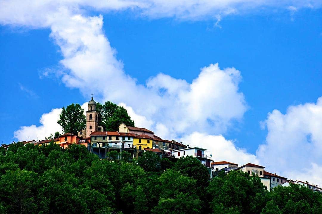 Pueblo italiano encaramado a una colina boscosa, con un campanario, bajo un cielo azul con nubes blancas.