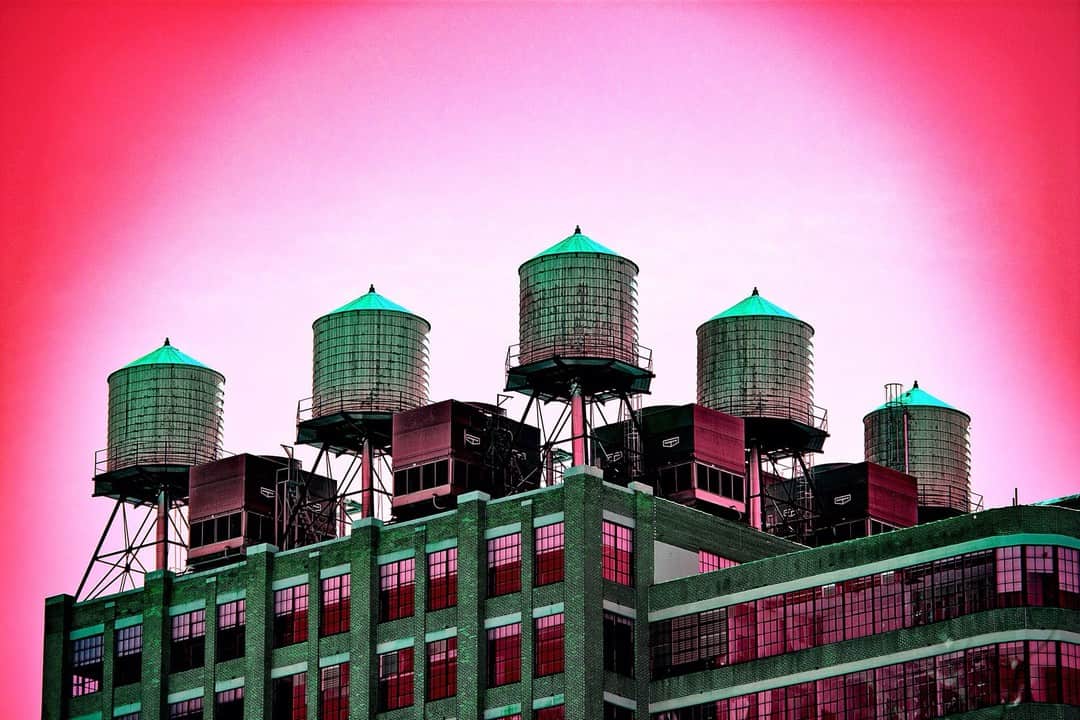 Cinco depósitos de agua sobre la azotea de un edificio industrial, con el cielo virado a rosa intenso y la estructura a verde.
