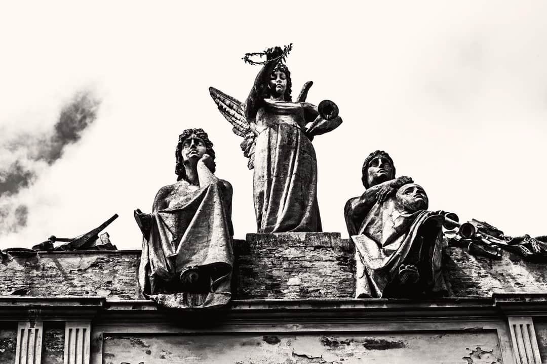 Grupo escultórico con un ángel sobre la cornisa de un edificio, recortado contra el cielo, en blanco y negro.