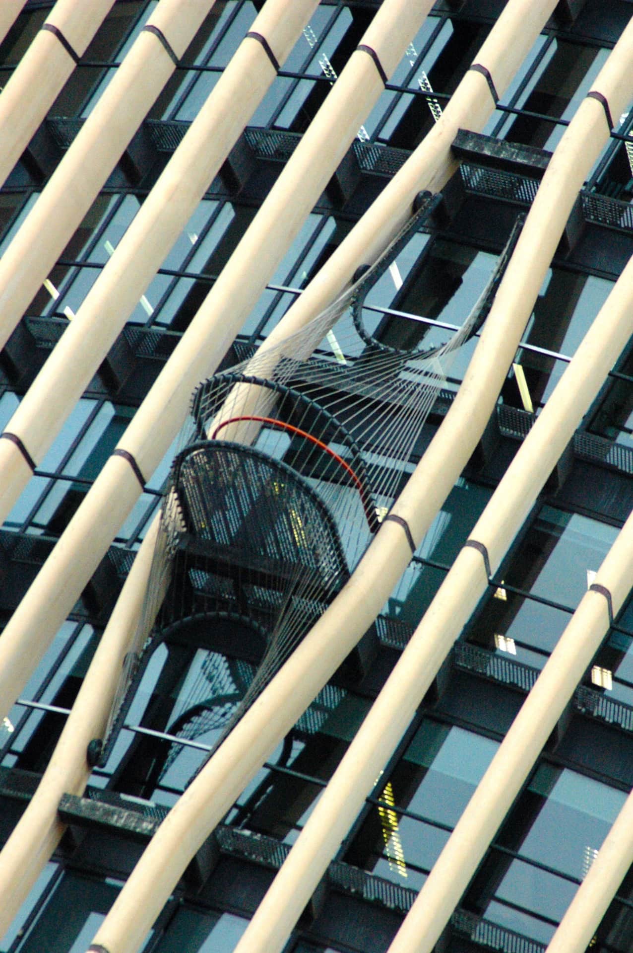 Tubos claros en diagonal cruzan el encuadre ante una torre acristalada con balcones y una red tensada con aro rojo.