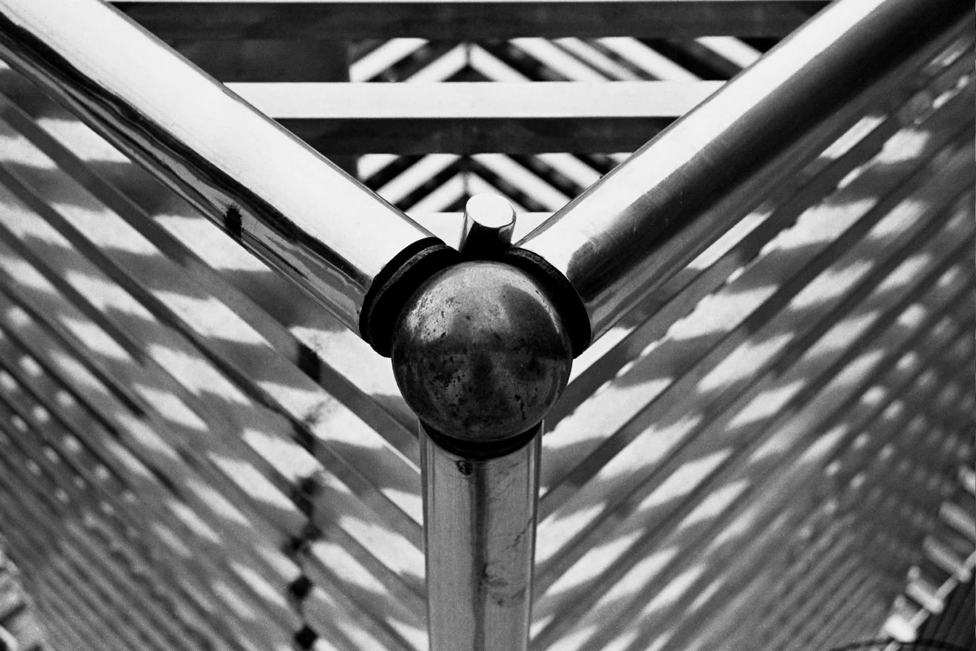Nudo esférico de acero enlaza tres tubos brillantes; detrás, una celosía de listones y sus sombras rayadas, en blanco y negro.