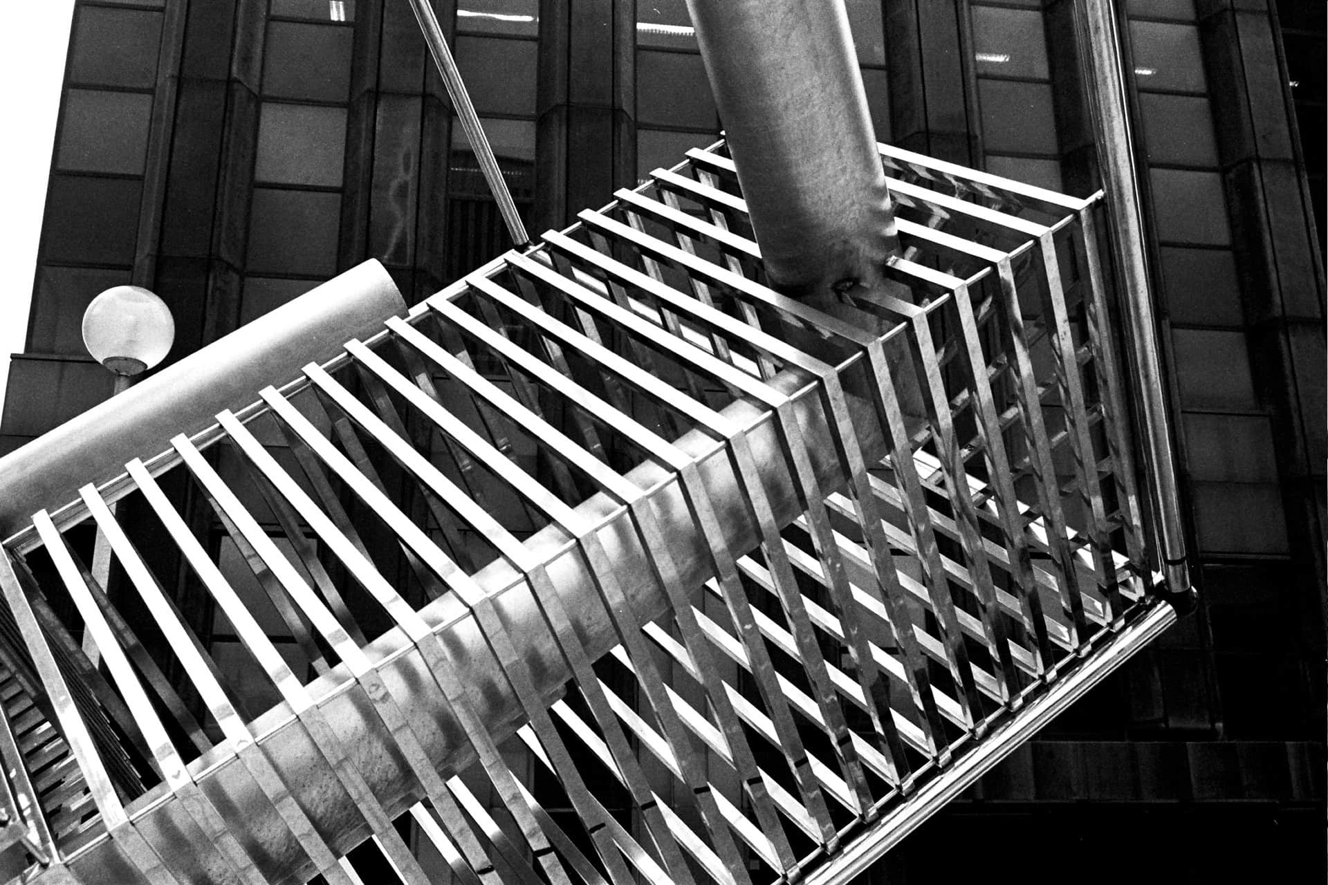 Estructura metálica de rejilla vista a contrapicado, en blanco y negro, con una farola de globo y un edificio de vidrio oscuro detrás.