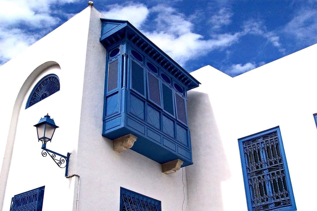 Mirador de madera azul en la esquina de una casa encalada, bajo un cielo azul con nubes.
