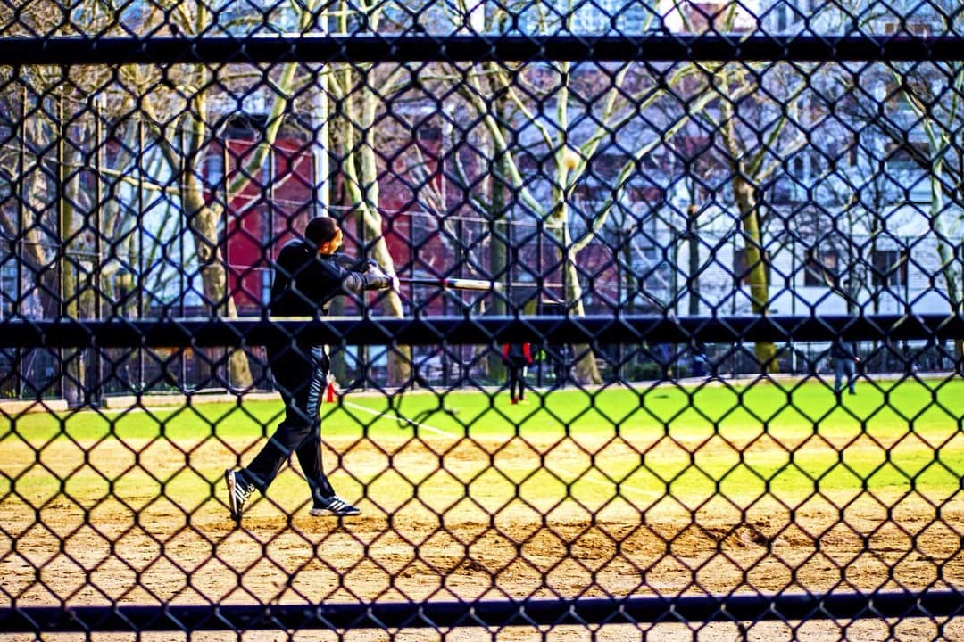 Jugador de béisbol visto a través de una valla metálica, con el campo verde y una vidriera de colores al fondo.