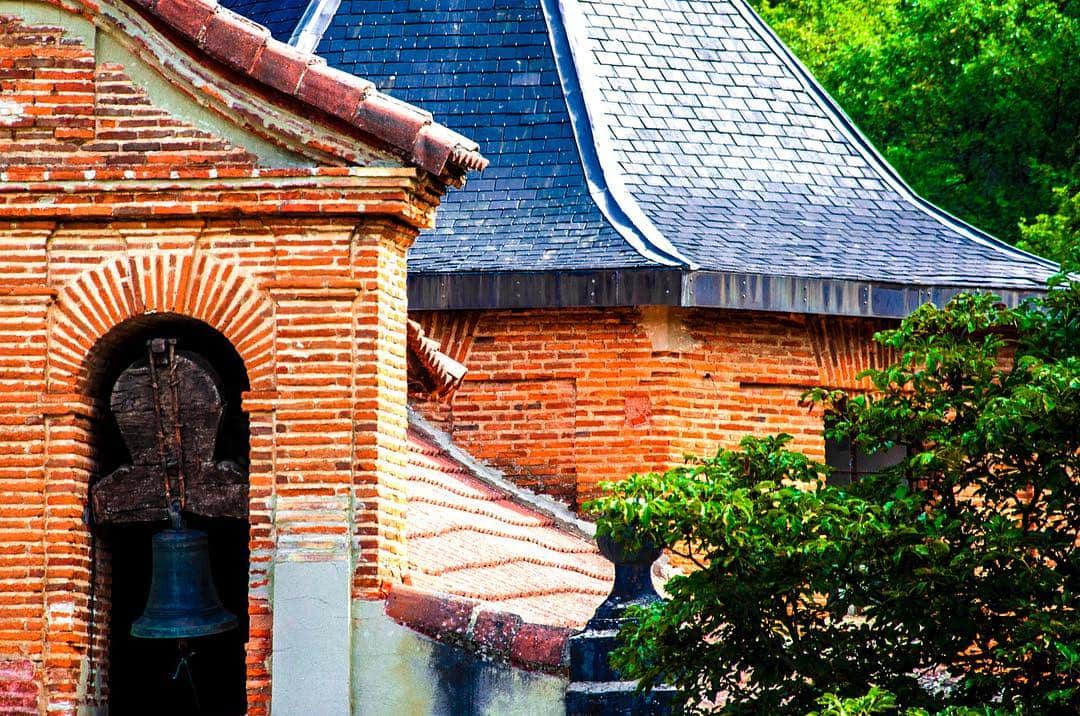 Campanario de ladrillo con una campana bajo un arco, junto a un tejado de pizarra azul y vegetación verde.