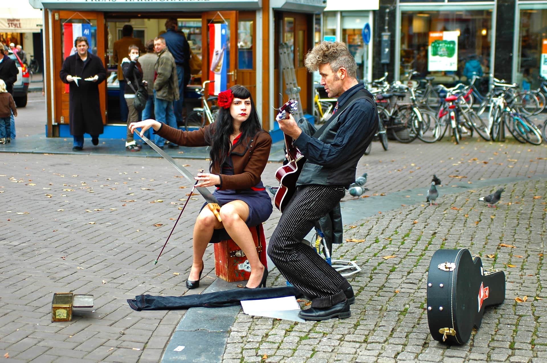 Dos músicos callejeros sobre adoquines: una mujer con una rosa roja en el pelo toca un serrucho con arco y un hombre, la guitarra.