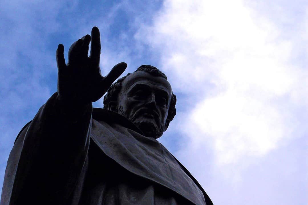 Estatua de una figura con la mano alzada en gesto de bendición, a contraluz contra un cielo nublado.