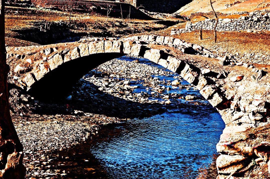 Puente de piedra de un arco sobre un arroyo de agua azul, con las orillas de grava.