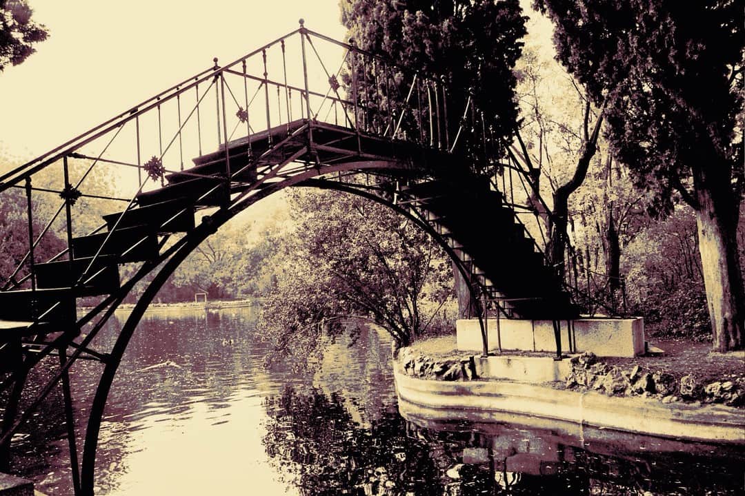 Pasarela metálica sobre un canal entre árboles, con la imagen virada a sepia y rosa.
