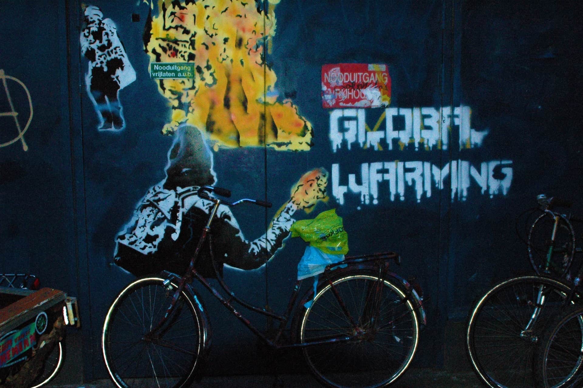 Bicicleta aparcada ante un grafiti de un encapuchado que parece pedalearla, junto al lema pintado Global Warming.