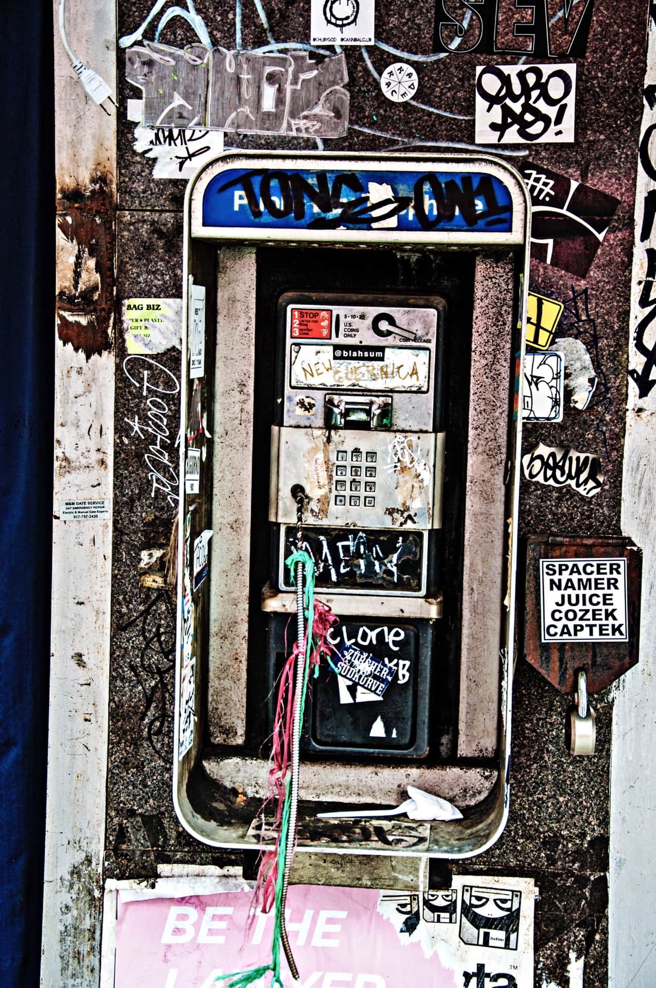 Teléfono público callejero cubierto de pegatinas y grafitis, con el cable colgando sin auricular y cintas de colores atadas.