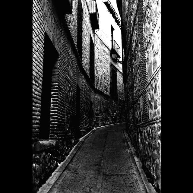 Callejón estrecho de Toledo en blanco y negro, entre muros de ladrillo y piedra, con el suelo empedrado brillante.