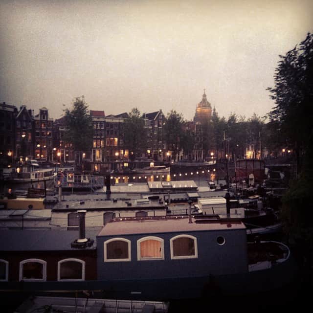Canal de Ámsterdam al anochecer, con casas flotantes amarradas en primer plano, las luces de la orilla encendidas y una cúpula recortada al fondo, en tonos sepia.