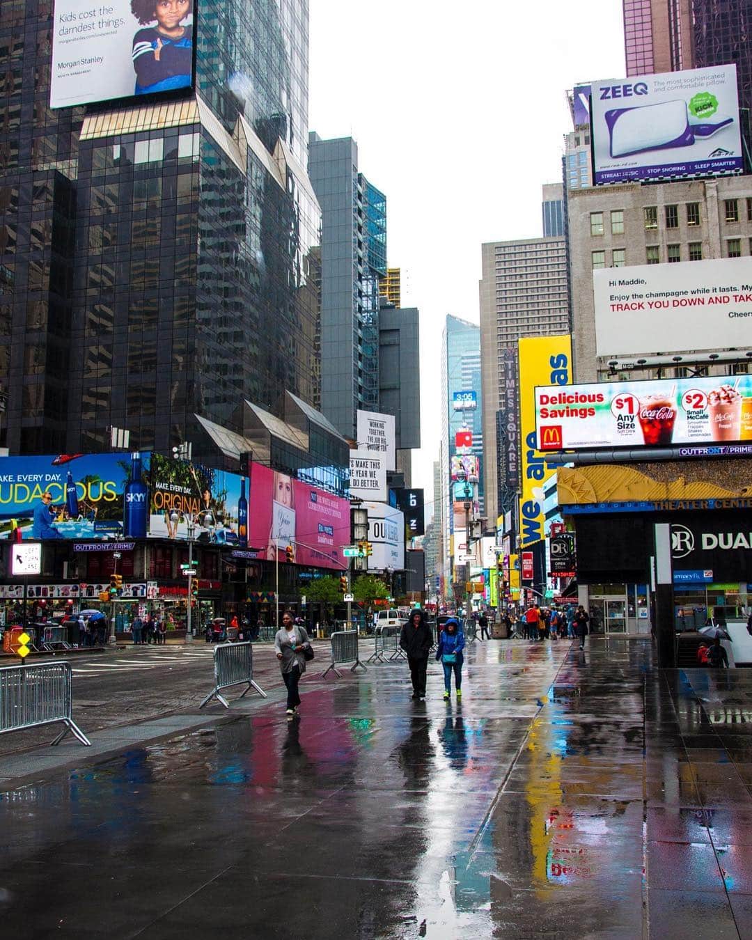 Times Square bajo la lluvia, con el pavimento mojado reflejando las pantallas publicitarias y unas pocas siluetas caminando.