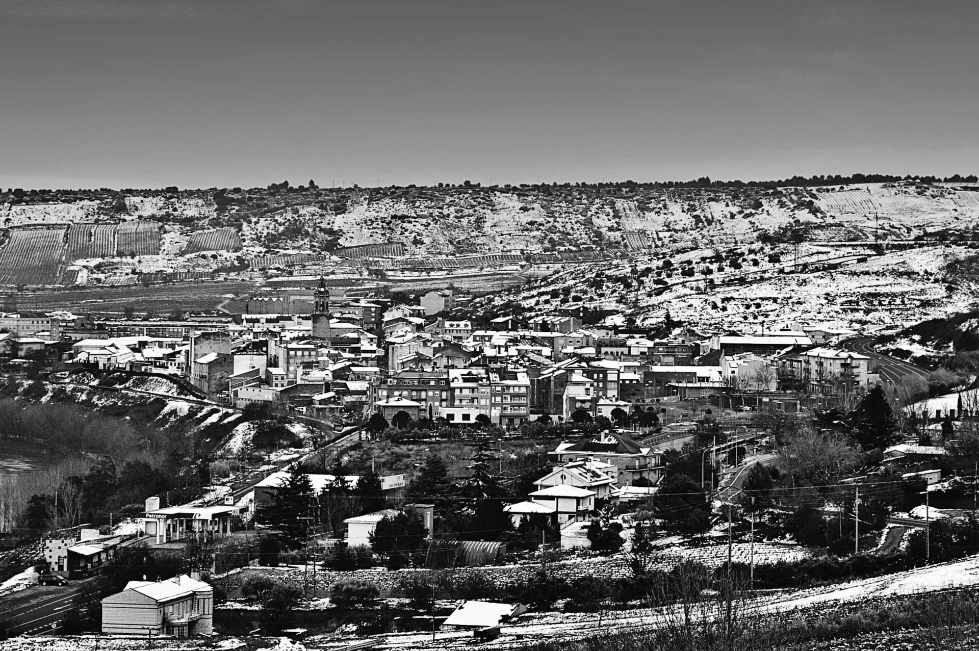 Pueblo nevado visto desde una loma, con la torre de la iglesia, una gasolinera abajo y laderas de viñedos, en blanco y negro.