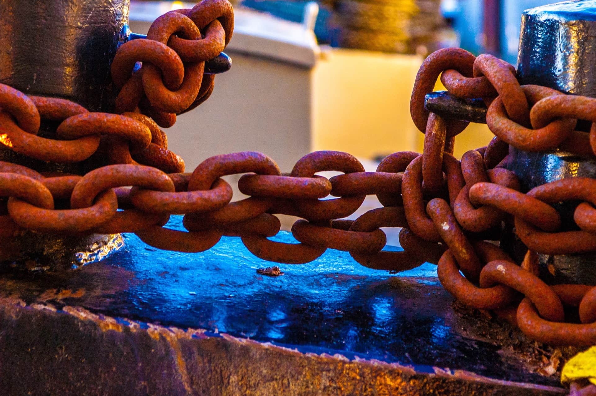 Primer plano de cadenas muy oxidadas tendidas entre dos postes metálicos sobre una superficie con reflejos azules y naranjas.