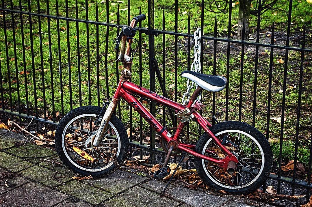 Bicicleta infantil roja apoyada y encadenada a una verja de hierro negra, sobre acera de adoquines y hojas caídas.