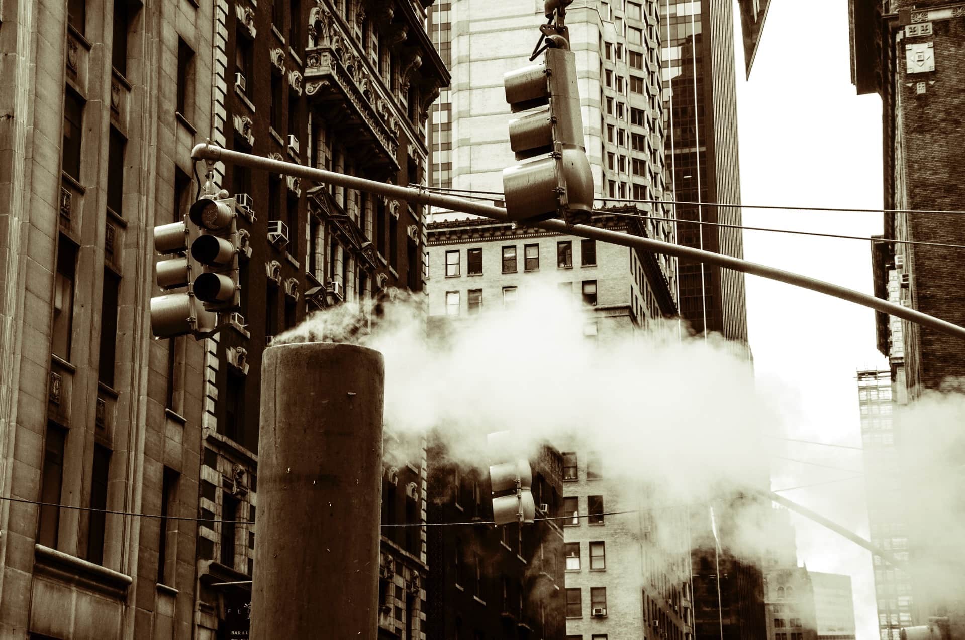 Chimenea de vapor humeando en mitad de la calle, entre semáforos suspendidos y fachadas de edificios altos, en virado sepia.