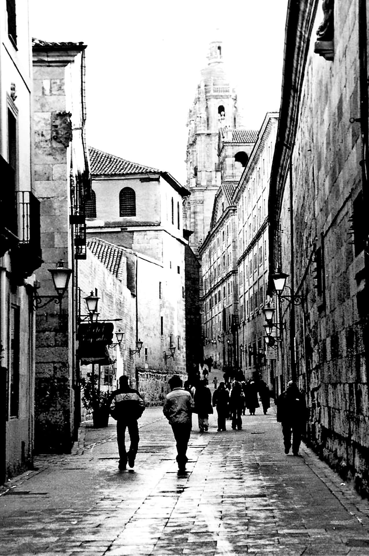 Calle estrecha y empedrada con viandantes de espaldas, faroles de forja en los muros y una torre barroca al fondo, en blanco y negro.
