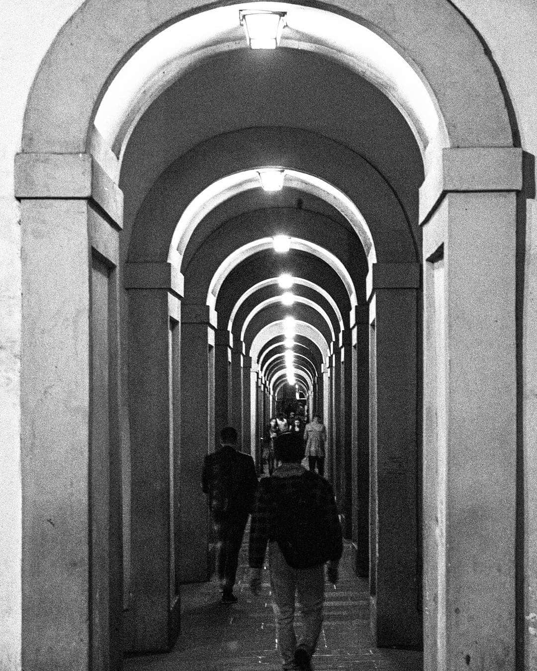 Galería abovedada larga con arcos de piedra y dos figuras caminando al fondo, en blanco y negro.