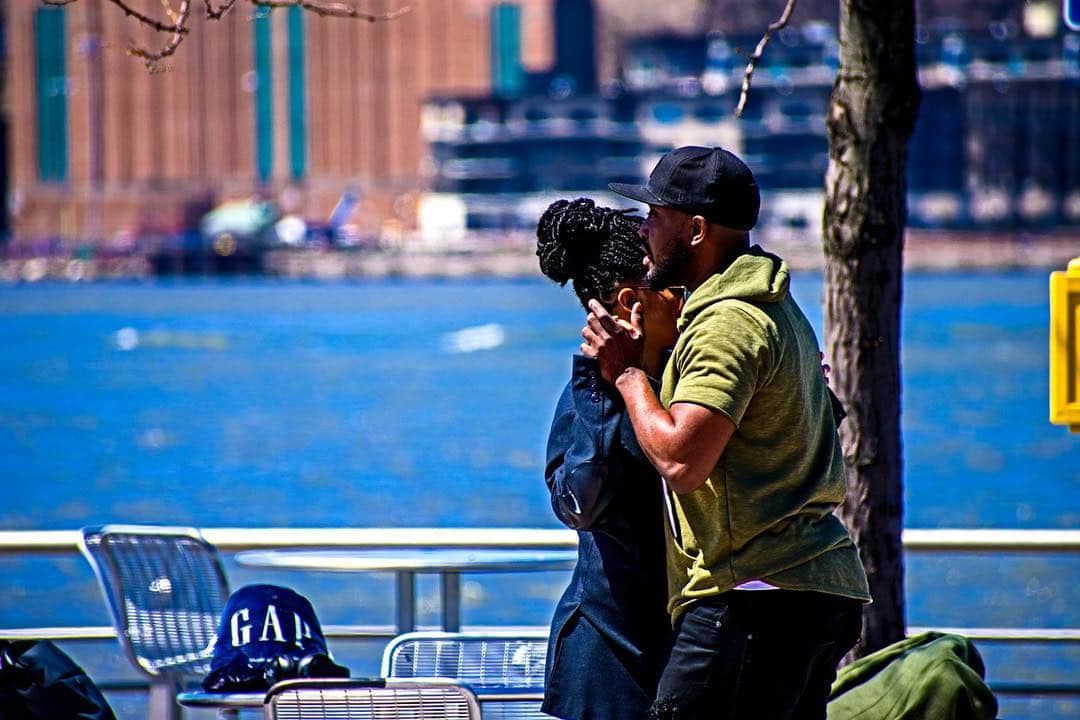 Pareja abrazada junto a la barandilla de un paseo marítimo, con el agua azul y el perfil de Manhattan al fondo.