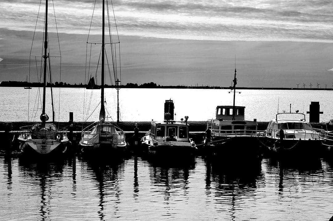 Barcos amarrados en un puerto, con los mástiles recortados sobre un cielo gris y el reflejo en el agua, en blanco y negro.