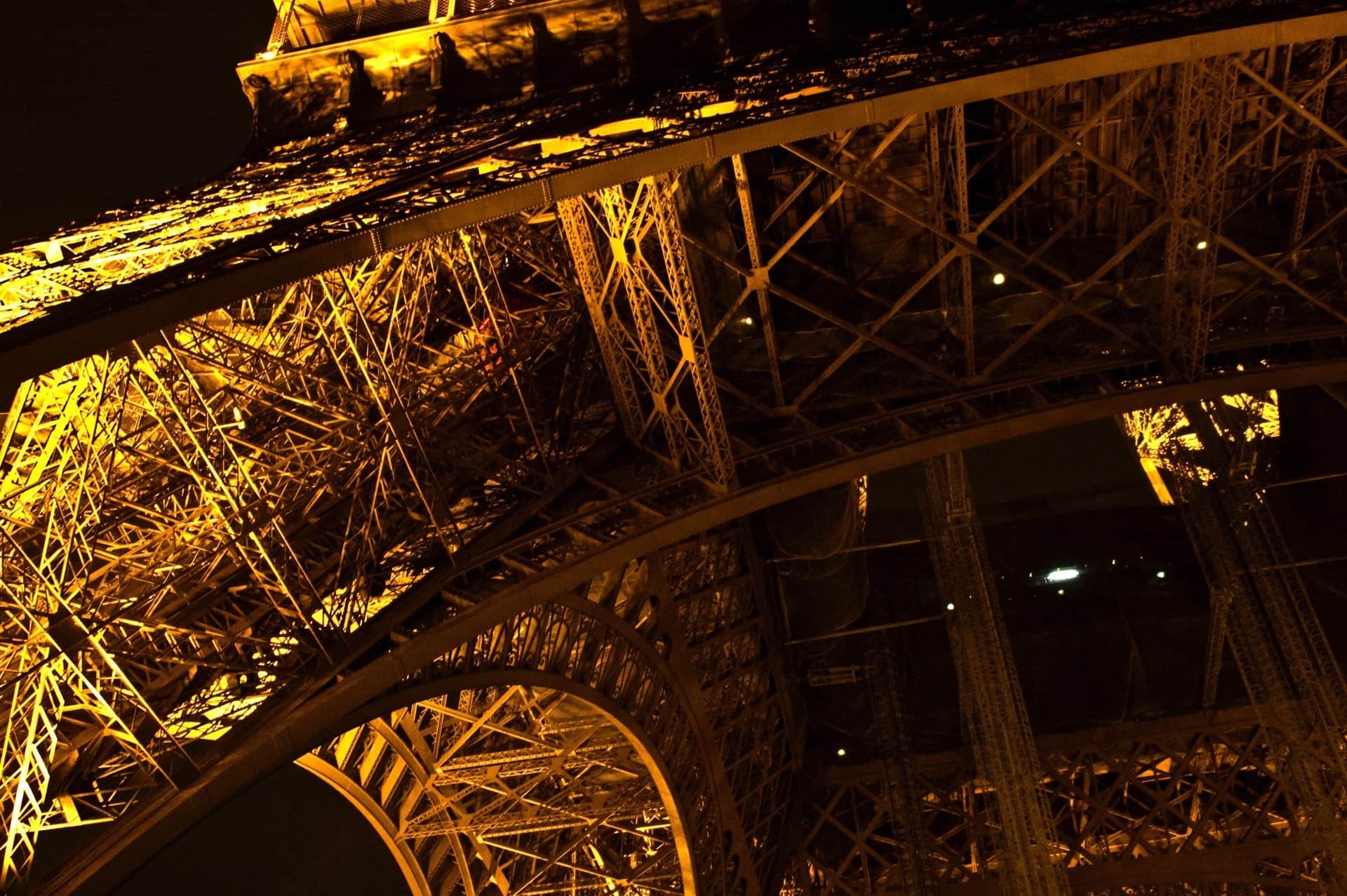 Estructura de hierro de la torre Eiffel en contrapicado, iluminada de noche, con el arco y las celosías en tonos dorados.
