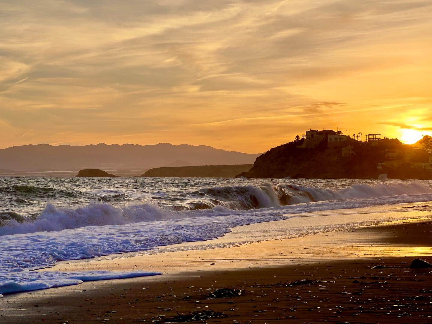 Playa al atardecer, con el sol bajo sobre el mar, un promontorio recortado a contraluz y la espuma de las olas en primer plano.