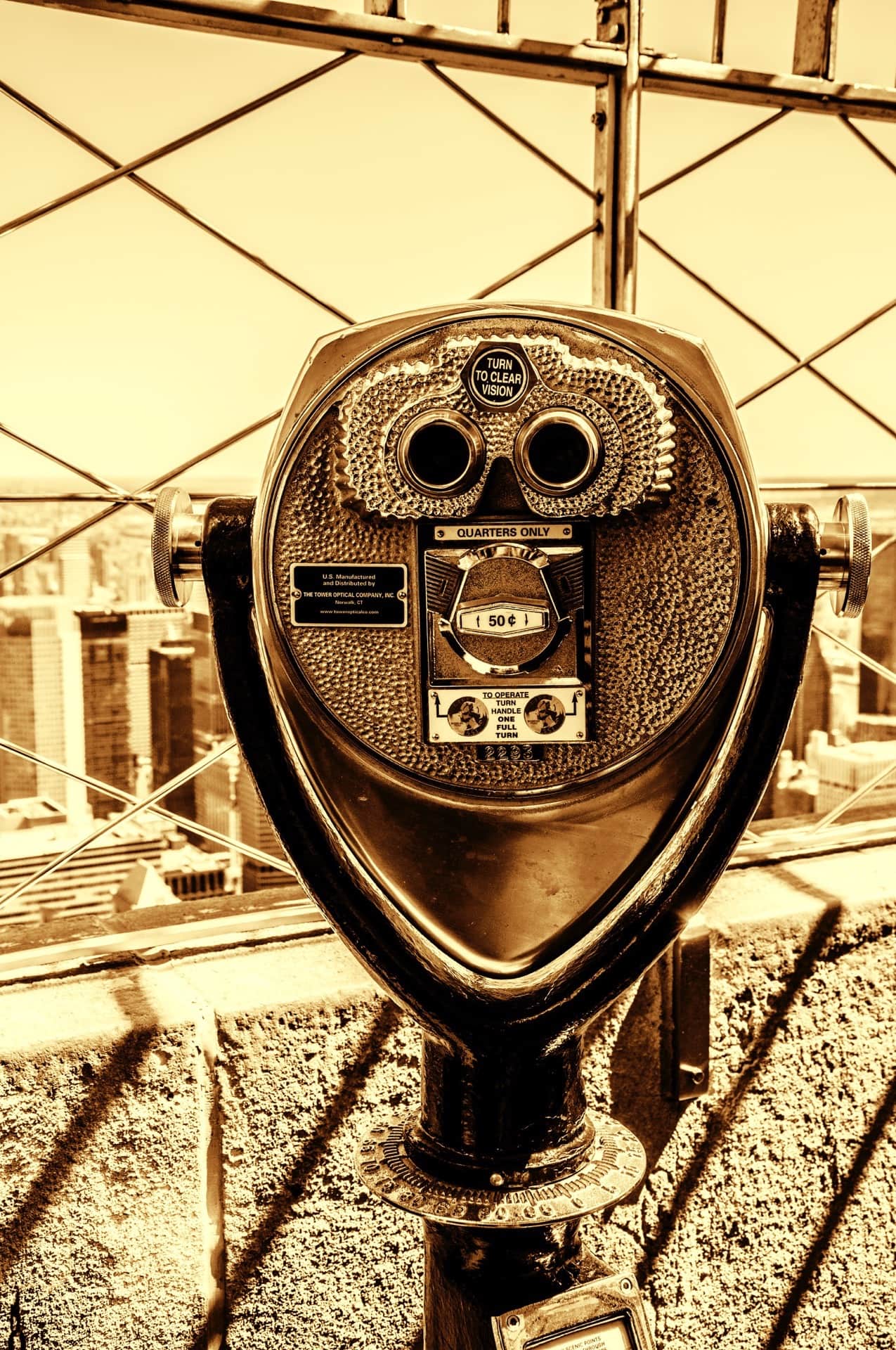 Visor binocular de monedas anclado al parapeto de un mirador en altura, virado en sepia, con rascacielos al fondo.