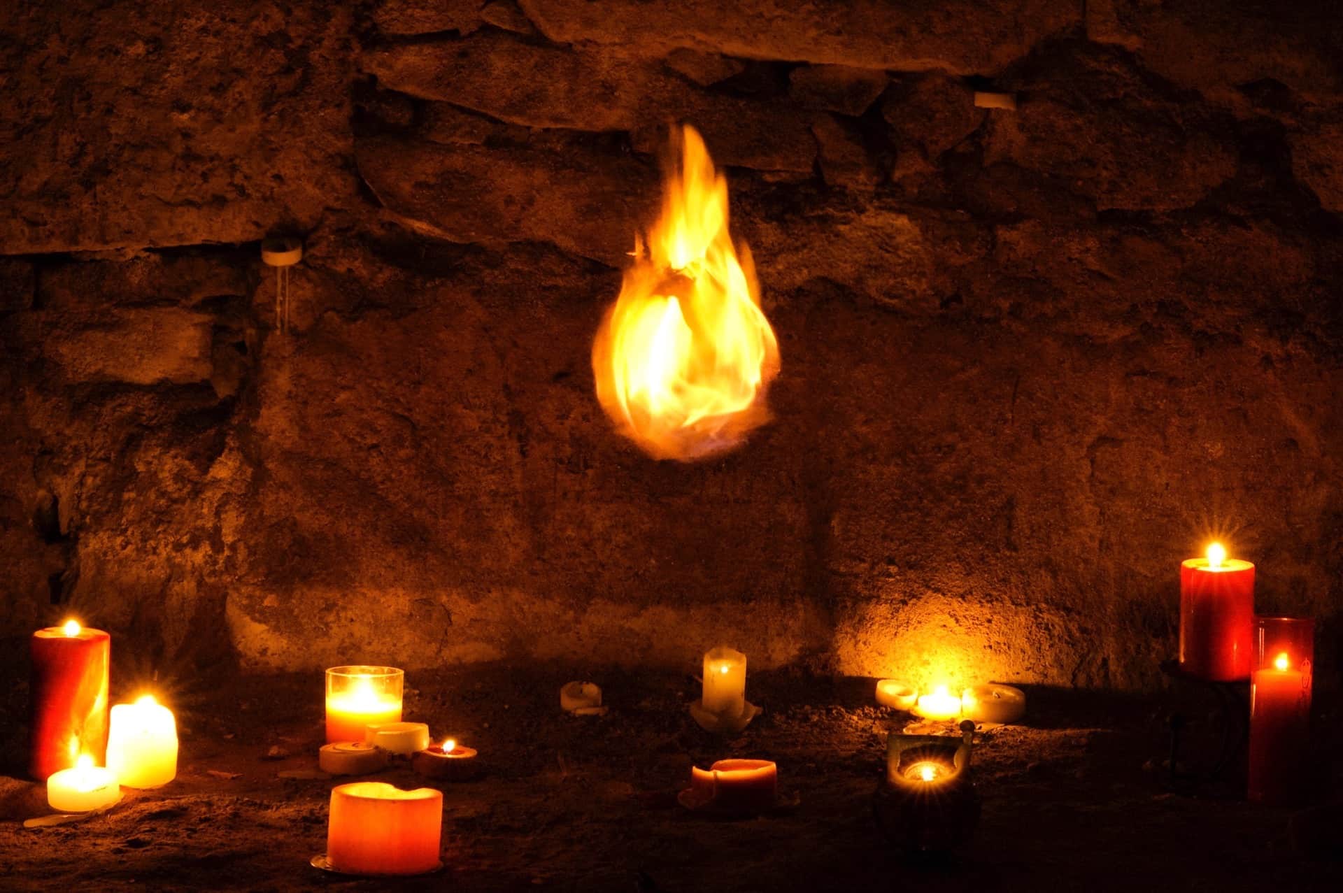 Llama suspendida en el aire ante un muro de piedra, con velas encendidas repartidas por el suelo a oscuras.