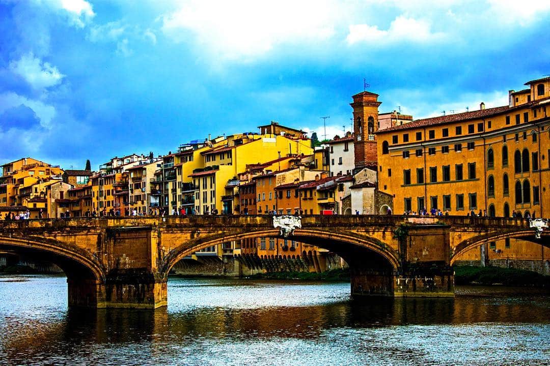 Puente sobre el Arno en Florencia, con las fachadas ocres reflejadas en el agua bajo un cielo de nubes.