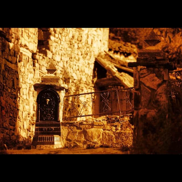 Fuente de piedra empotrada en un muro de sillares, con una reja de hierro y una escalera al fondo, todo virado en tonos sepia y anaranjados.