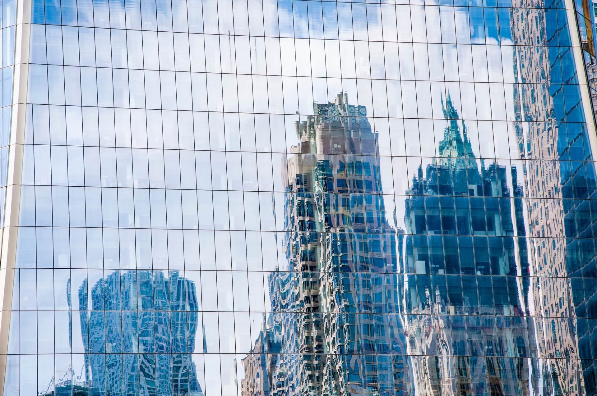 Fachada acristalada de un rascacielos que refleja, deformadas, las torres vecinas y el cielo con nubes.