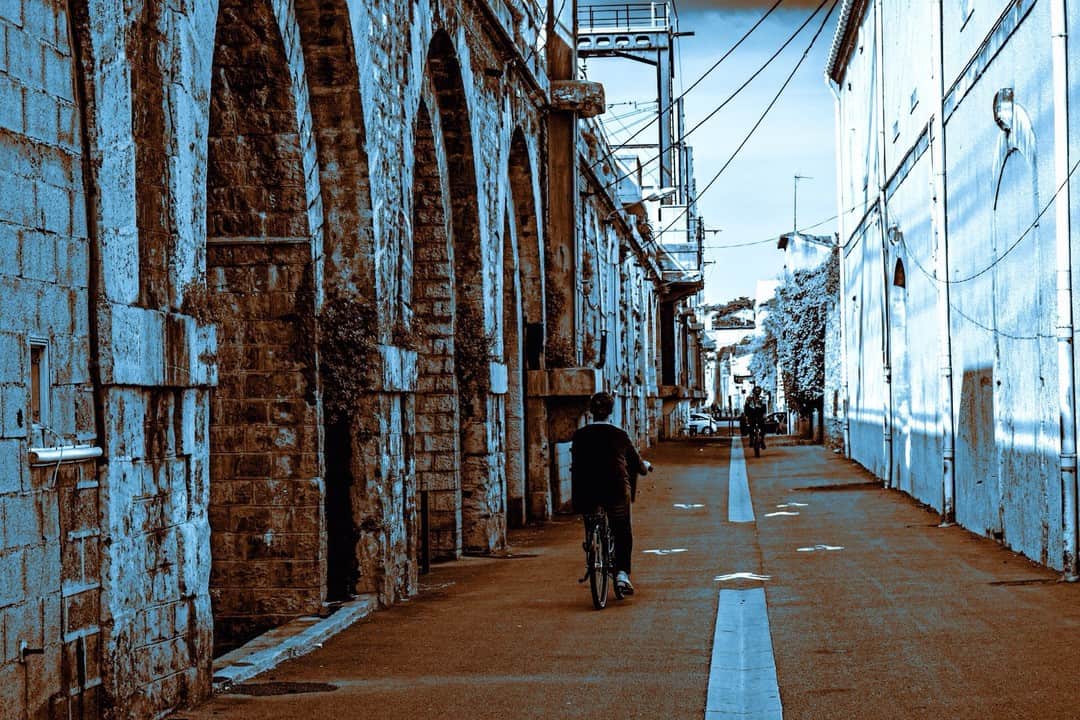 Calle larga con soportales de piedra a un lado y un mural azul al otro, con un ciclista al fondo.