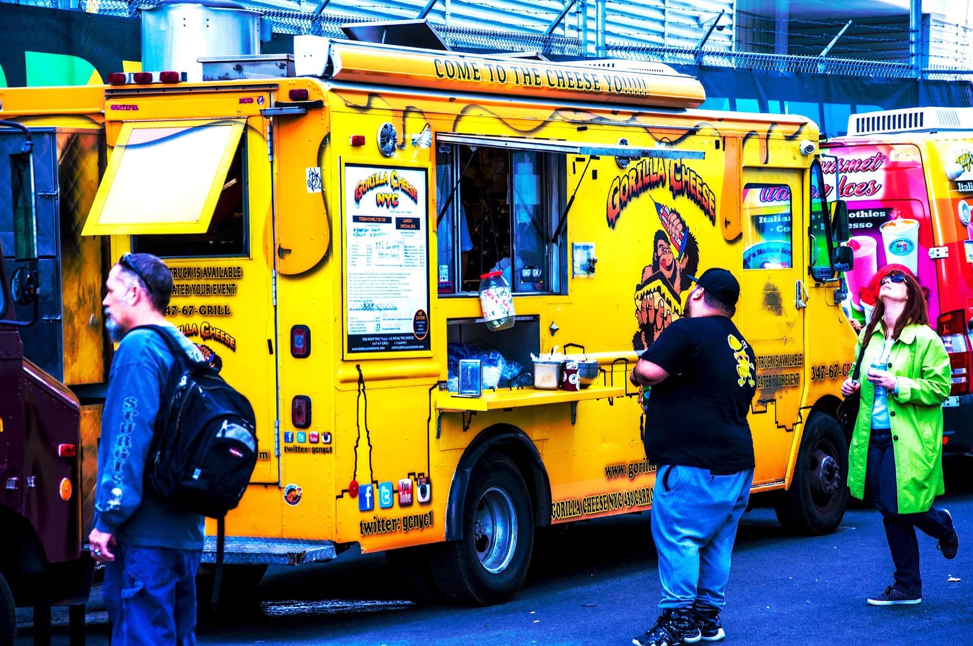 Camión de comida amarillo rotulado Gorilla Cheese NYC, con un gorila pintado y varias personas alrededor, en color saturado.