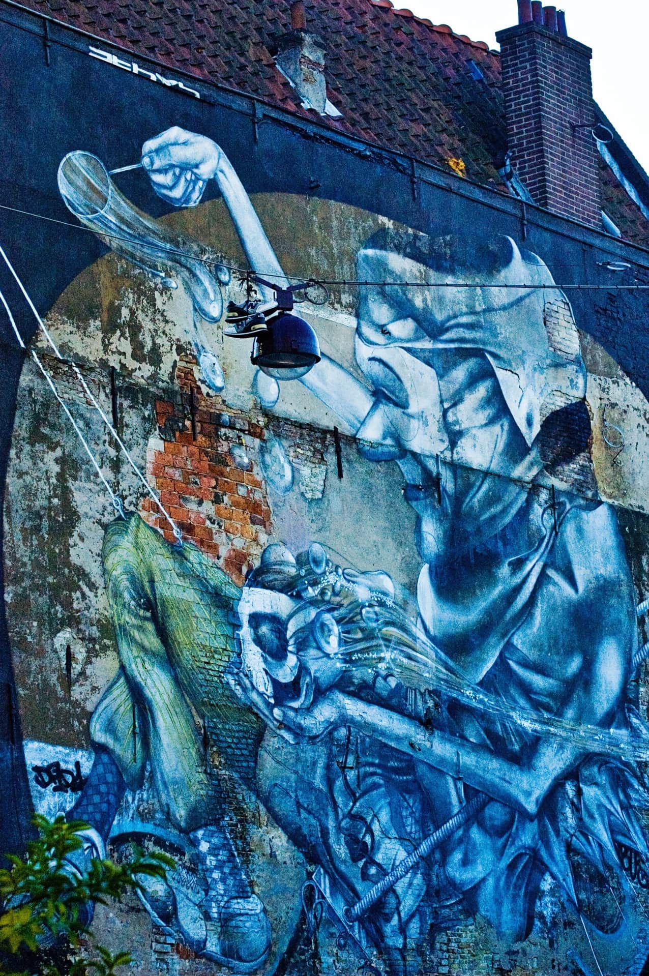 Mural de dos figuras gigantes en tonos azules sobre la medianera de una casa con tejado de teja roja.