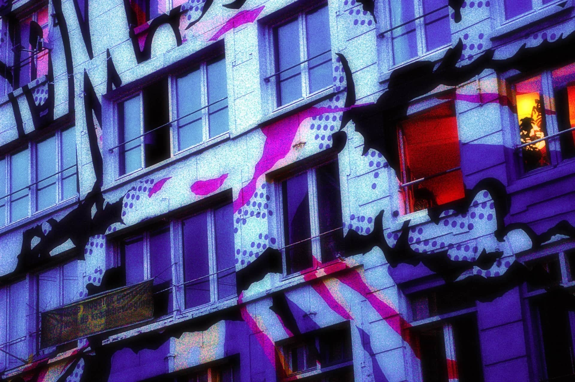 Fachada de viviendas cubierta por un mural de manchas negras y lunares, con una persiana roja, virada a violeta y magenta.