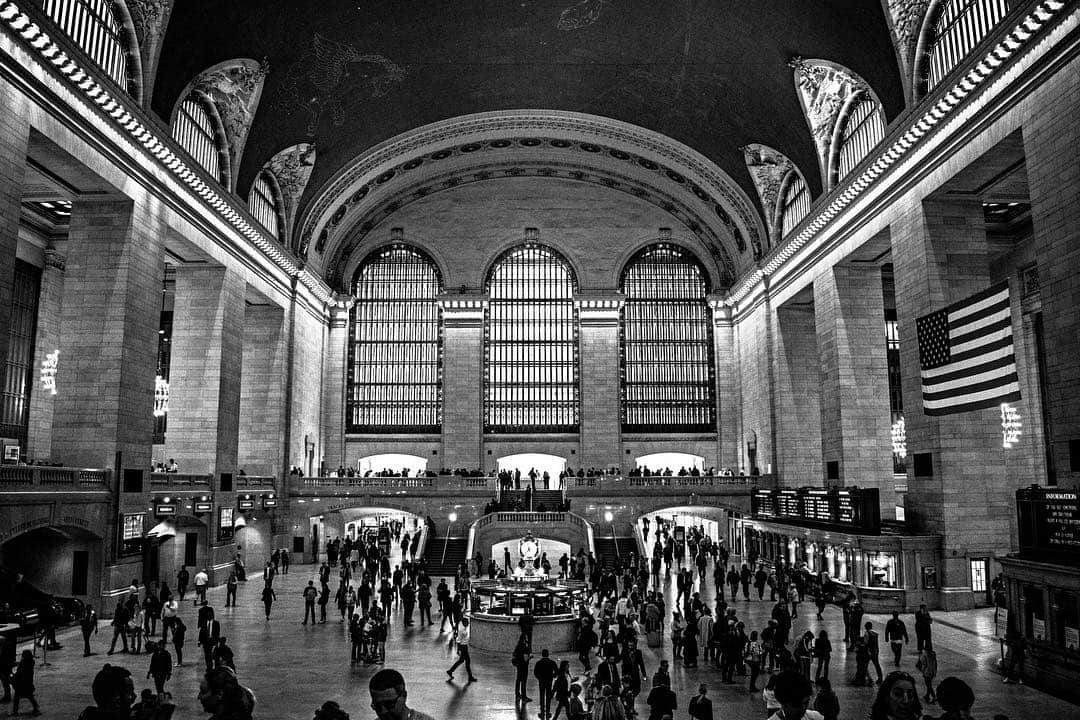 Vestíbulo principal de Grand Central lleno de gente, con los grandes ventanales al fondo y una bandera colgada, en blanco y negro.